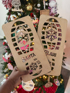 Wooden Santa footprint stencils spelling SANTA and CLAUS held up in front of a decorated Christmas tree