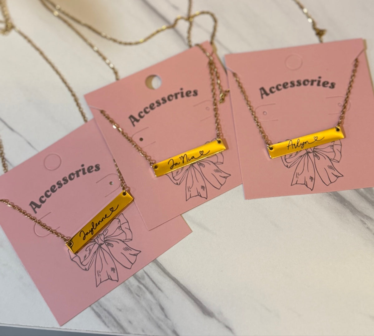 Three gold engraved name bar necklaces personalized with Jaylenne, Ja'Nia and Arlyn on pink accessories display cards